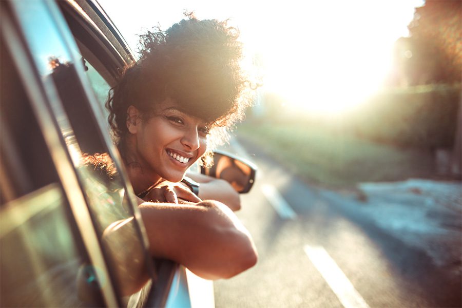Client Center - Closeup Portrait of a Smiling Young Woman Sticking Her Head Out the Window While on a Road Trip on a Sunny Day